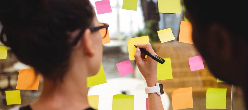 Uma pessoa escrevendo em um post-it colado em um quadro de vidro