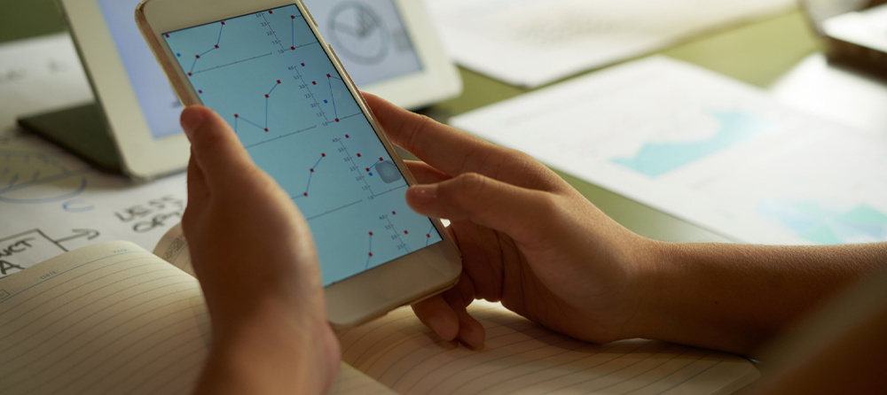 Imagem de duas mãos segurando um celular com gráficos na tela