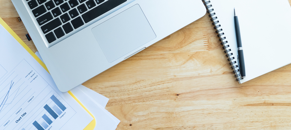 Imagem de um notebook com documentações impressas ao lado e uma agenda, em cima de uma mesa.