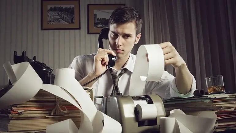 AN ACCOUNTANT ON THE PHONE, SURROUNDED BY PILES OF PAPERWORK THAT HE IS REVIEWING. 