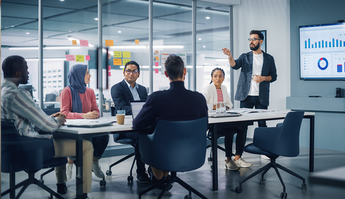 Imagem de uma sala de reuniões, com diversos colaboradores da empresa sentados, ouvindo uma apresentação de indicadores  