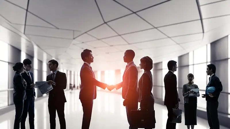 TWO BUSINESS MEN SHAKING HANDS IN A HALLWAY WITH OTHER PEOPLE. 
