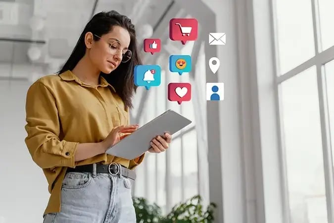 Na imagem, há uma mulher utilizando óculos de grau e vestindo uma blusa amarela. Ela segura um ipad. Acima dele surgem ícones flutuantes que simbolizam elementos onde o marketing atua, como: coração que remete ao like das redes sociais, sino que remete as notificações, pin que remete a localização, carrinho de supermercado que remete à compras, e uma carta que simboliza o envio de e-mails. 